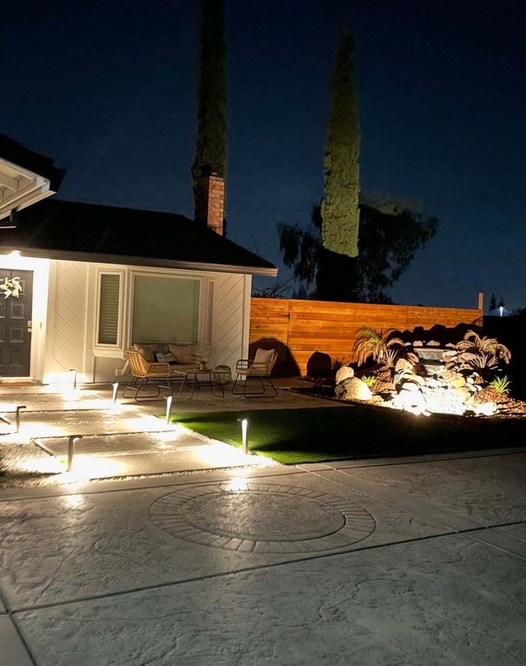 Nighttime backyard patio with illuminated concrete, modern house, wooden fence, and landscaped garden with cypress trees