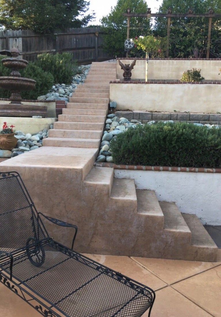 Backyard landscaping with tiered brick steps, retaining walls, decorative rocks, shrubs, and wooden fence with trees in background