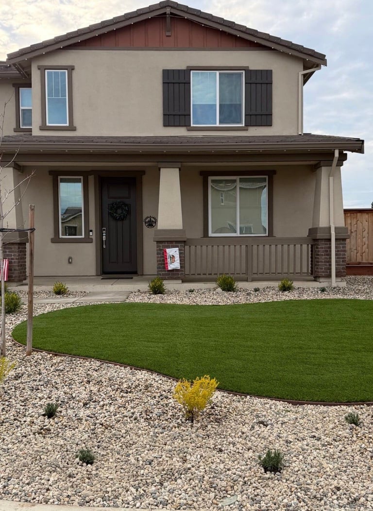Modern two-story house with beige exterior, dark shutters, front porch, manicured lawn, and gravel landscaping with plants